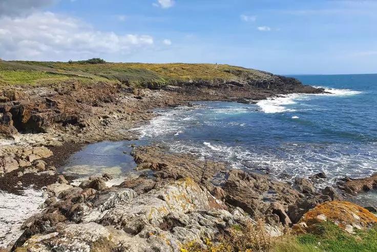 Felsige K&uuml;stenlinie mit Wellen, kleiner Bucht, grasbewachsene Klippen und blauem Himmel mit vereinzelten Wolken