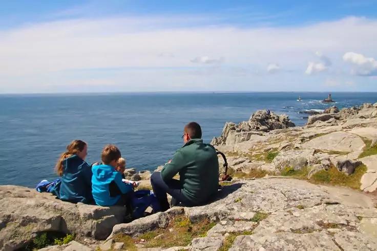 Drei Personen sitzen auf Felsen und blicken aufs Meer; zwei tragen blaue Jacken, eine eine gr&uuml;ne Jacke. Felsenk&uuml;ste, Meer, wolkiger Himmel und ein Leuchtturm im Hintergrund