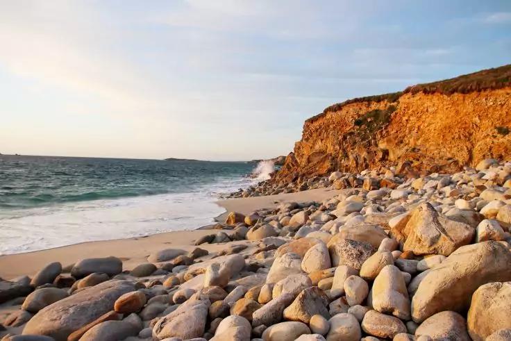 Strand mit glatten, runden Felsen im Vordergrund, schmaler Sandstreifen, Meereswellen, orange-braune Felsklippe rechts, heller Himmel mit d&uuml;nnen Wolken