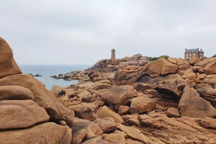 R&ouml;tlich-braune Felsformationen an der K&uuml;ste, im Hintergrund ein Leuchtturm auf einem Felsvorsprung, ein Geb&auml;ude rechts, Meer links und bedeckter Himmel