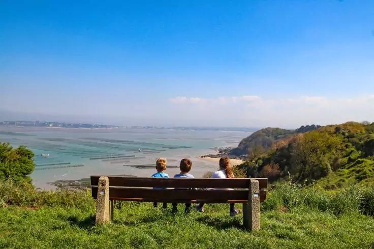 Drei Personen sitzen mit dem R&uuml;cken zur Kamera auf einer Parkbank auf einer grasbewachsenen Anh&ouml;he und blicken auf das Meer und die K&uuml;ste