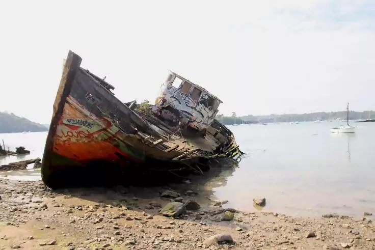 Rostiges, besch&auml;digtes Schiff liegt schr&auml;g auf einem kiesigen Ufer; ruhiges Wasser, Segelboot und entfernte Uferlinie im Hintergrund