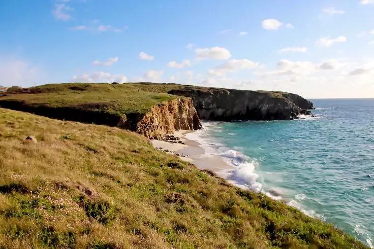 Grasbewachsene Klippe mit kleinem Sandstrand, Wellen und blauem Meer unter wolkigem Himmel