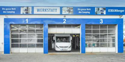 Blaue Werkstatt mit drei nummerierten Garagentoren (1-3), in der Mitte ein wei&szlig;es Wohnmobil halb in der Einfahrt, eine Person im Fahrerhaus, Schriftzug 'WERKSTATT' und Werbebanner an der Fassade