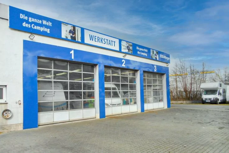 Gebäude mit drei blauen Garagentoren nummeriert 1–3, oben Schild 'WERKSTATT' und 'Die ganze Welt des Camping', gepflasterter Vorplatz, rechts geparktes weißes Wohnmobil, teilweise bewölkter Himmel