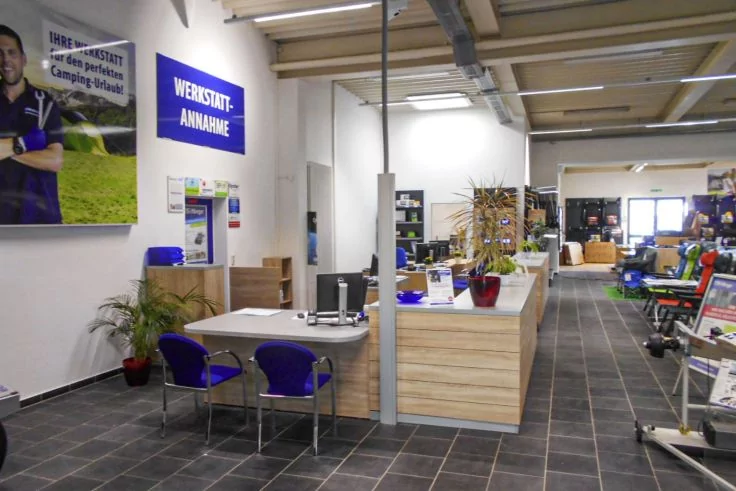 Innenraum mit hölzerner Empfangstheke, Schreibtisch mit zwei blauen Stühlen, Topfpflanzen, Regale, Poster an der Wand mit dem Schriftzug 'WERKSTATT-ANNAHME', gefliester Boden, Deckenlampen