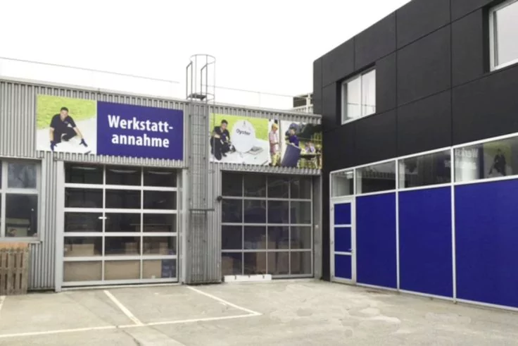 Außenansicht einer Werkstatt mit zwei großen gläsernen Rolltoren und einem Schild 'Werkstattannahme' über dem linken Tor; graue Blechfassade, blaue Paneele rechts, leerer Betonvorplatz