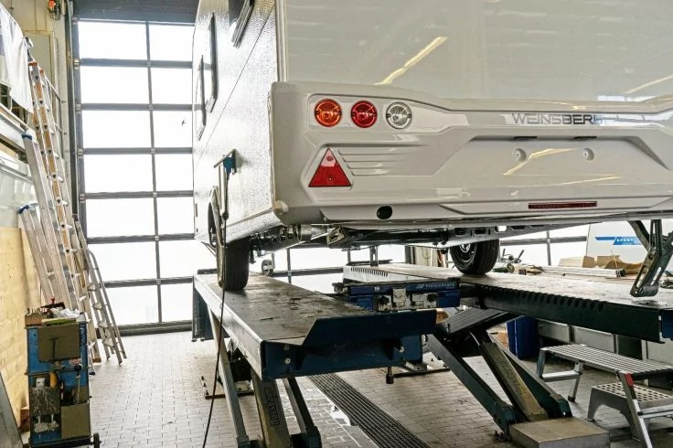 Heck eines Wohnwagens auf einer Hebebühne in einer Werkstatt; Rücklichter, rotes Dreieck, Rad, Metallbühnen, Leiter an der Wand, großes Garagentor mit Fenstern