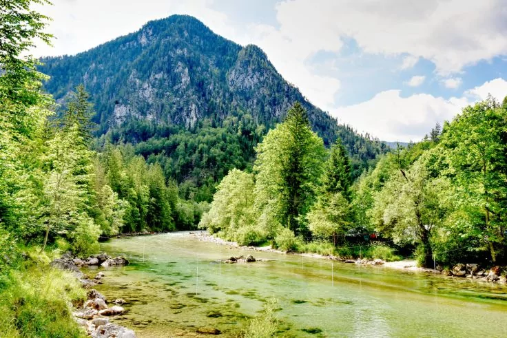 Fluss mit klarem Wasser, sichtbarem Flussbett und Steinen, baumbewachsene Ufer und bewaldeter Berg im Hintergrund unter teils bew&ouml;lktem Himmel
