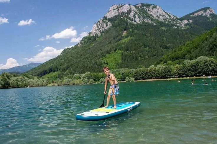 Person steht auf einem Stand-up-Paddleboard auf klarem See, bewaldete Berge im Hintergrund, blauer Himmel mit wenigen Wolken
