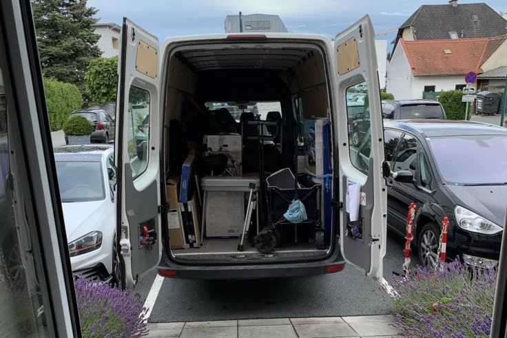 Ge&ouml;ffneter wei&szlig;er Lieferwagen von hinten, Laderaum mit Kartons und Ausr&uuml;stung, an einem Gegenstand h&auml;ngt eine blaue Einwegmaske, links und rechts geparkte Autos, lila bl&uuml;hende Pflanzen am Eingangsweg, Wohnh&auml;user und Stra&szlig;e im Hintergrund