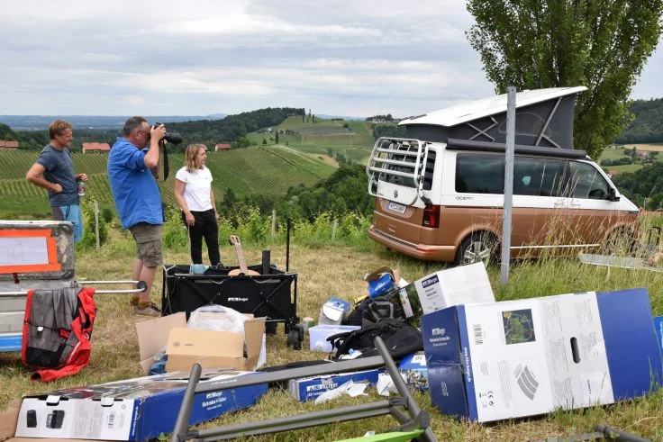 Drei Personen neben einem Camper mit aufgeklapptem Dach; Kartons und Ausr&uuml;stung im Vordergrund, Weinberge und H&uuml;gel im Hintergrund, bew&ouml;lkter Himmel