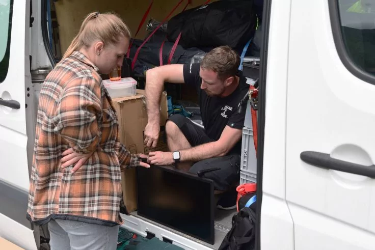 Wei&szlig;er Kastenwagen mit ge&ouml;ffneter Seitent&uuml;r; eine Frau in karierter Jacke steht am Einstieg, ein Mann im schwarzen T-Shirt kniet im Laderaum und fasst an einen Flachbildschirm; Kartons und Taschen im Laderaum sichtbar
