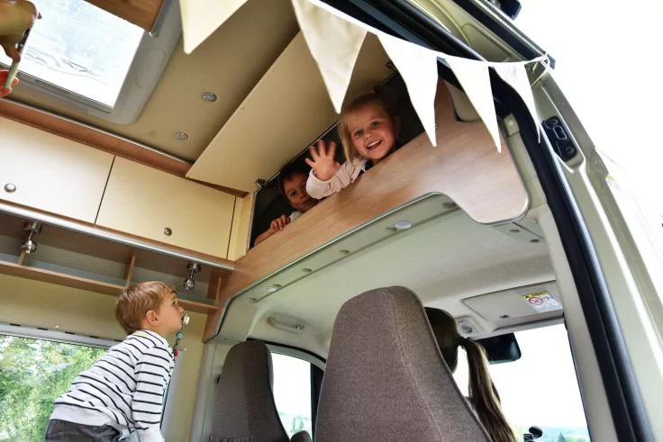 Innenraum eines Campers mit zwei Kindern: eines in einem Hochbett winkt, das andere steht unten und schaut nach oben; Wimpel, Schr&auml;nke und Vordersitze sichtbar