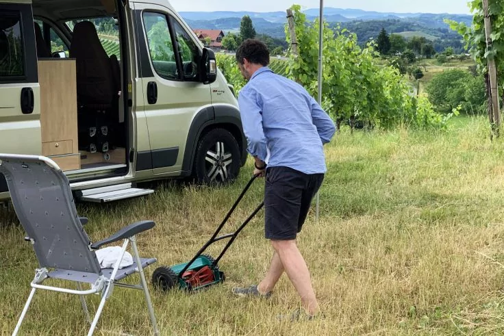 Eine Person in blauem Hemd und dunklen Shorts schiebt einen Rasenm&auml;her auf einer Wiese neben einem geparkten Campervan; Klappstuhl links; Reihen von Pflanzen und H&uuml;gel im Hintergrund