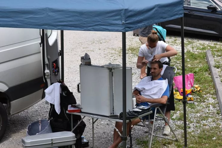 Unter einem blauen Pavillon sitzt eine Person auf einem Klappstuhl mit einem Tuch um die Schultern, eine zweite Person steht hinter ihr und ber&uuml;hrt ihren Kopf; daneben ein Tisch mit offenem Spiegelkoffer und ein wei&szlig;er Lieferwagen auf Kies