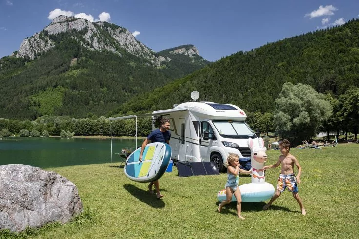 Drei Personen mit aufblasbaren Schwimmringen und einem Stand-up-Paddle neben einem wei&szlig;en Wohnmobil auf einer gr&uuml;nen Uferwiese; im Hintergrund See, Wald, Berge und blauer Himmel mit Wolken