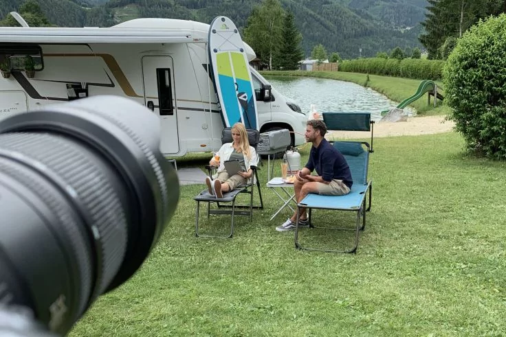 Gro&szlig;es Kameraobjektiv im Vordergrund links; zwei Personen sitzen auf Liegest&uuml;hlen vor einem Wohnmobil auf einer Wiese; Surfboard lehnt am Wohnmobil; See mit Rutsche und bewaldete Berge im Hintergrund
