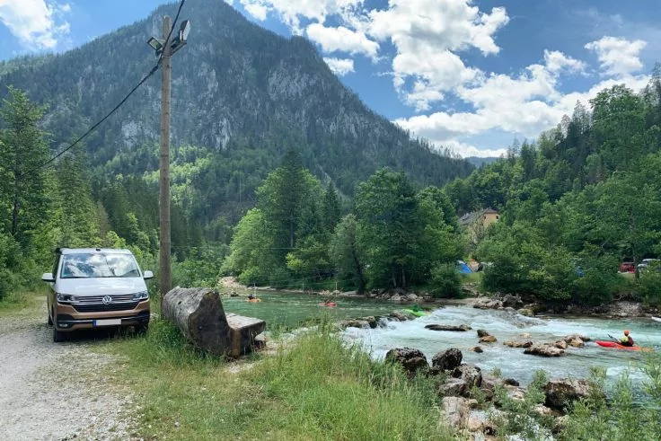 Wei&szlig;er Transporter links auf Schotterweg, h&ouml;lzerne Bank neben gro&szlig;em Fels und Strommast; grasiges Flussufer, t&uuml;rkisfarbener Fluss mit Felsen und orangem Kajak mit Person; bewaldete H&uuml;gel und Berg im Hintergrund, teils wolkiger Himmel