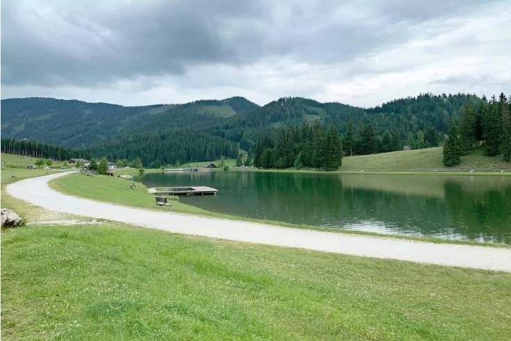 Grasufer mit geschwungenem Kiesweg, kleiner Holzsteg am See, bewaldete H&uuml;gel und bew&ouml;lkter Himmel