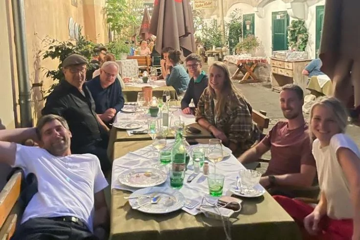 Mehrere Personen sitzen an einem langen, gedeckten Au&szlig;entisch in einem beleuchteten Restaurantbereich; auf dem Tisch Teller, Gl&auml;ser und gr&uuml;ne Flaschen; Sonnenschirme, Pflanzen und Steinw&auml;nde im Hintergrund