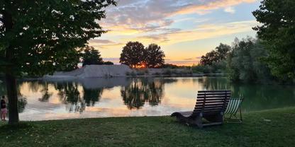 Grasufer mit zwei Liegest&uuml;hlen, ruhiger See mit Spiegelung von B&auml;umen, kleiner Sandh&uuml;gel in Bildmitte, Himmel mit orange-blauen Wolken am Horizont