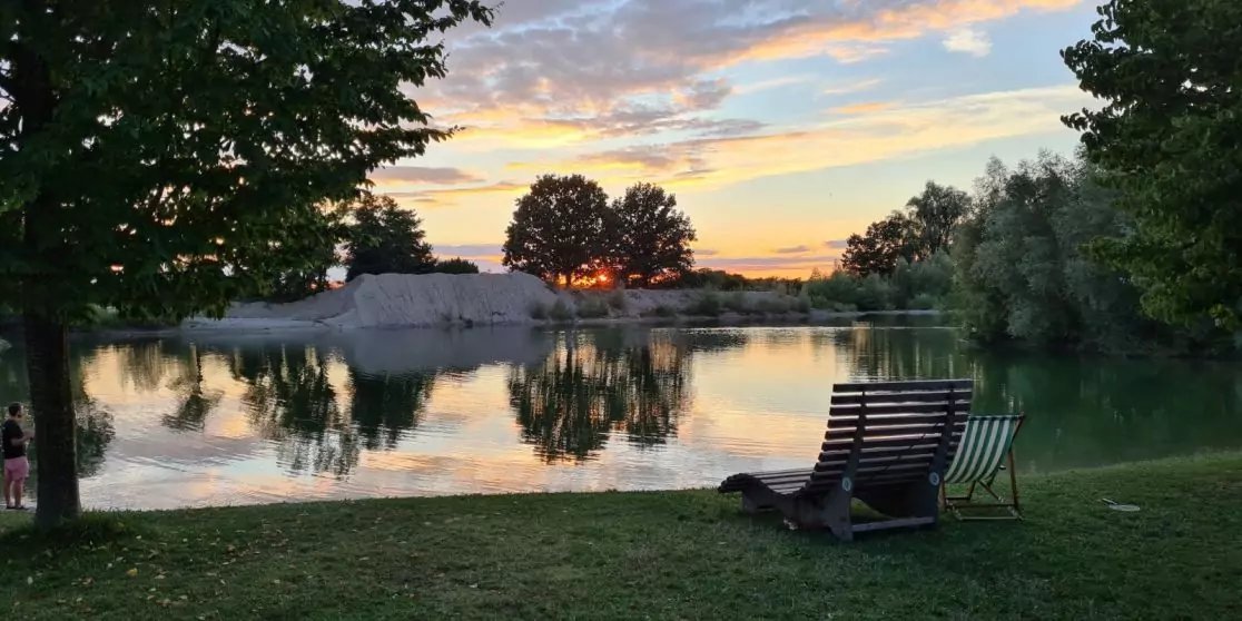Grasufer mit zwei Liegest&uuml;hlen, ruhiger See mit Spiegelung von B&auml;umen, kleiner Sandh&uuml;gel in Bildmitte, Himmel mit orange-blauen Wolken am Horizont