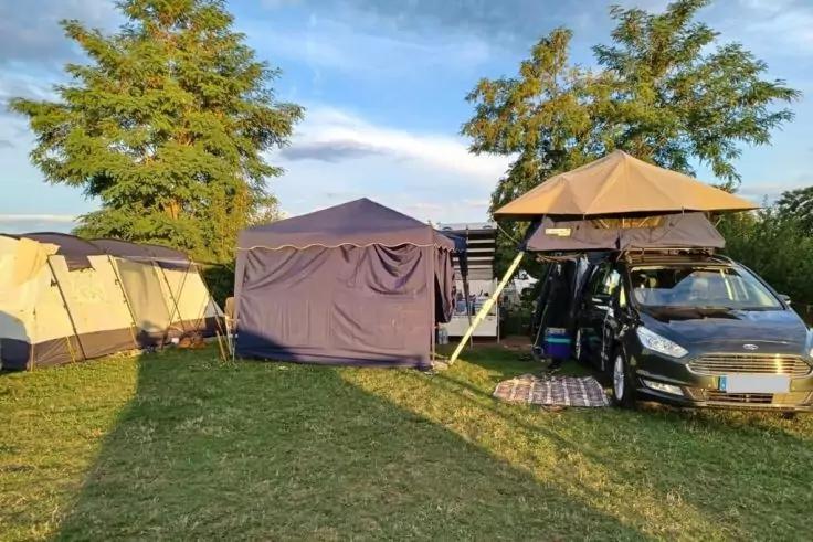 Campingplatz mit mehreren Zelten auf einer Wiese, Auto mit ge&ouml;ffnetem Dachzelt rechts, B&auml;ume und blauer Himmel im Hintergrund