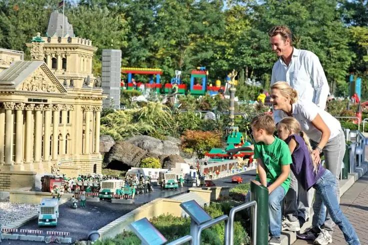 Zwei Erwachsene und zwei Kinder lehnen an einem Gel&auml;nder und betrachten im Freien Miniaturgeb&auml;ude und -fahrzeuge mit einem Wasserbecken davor