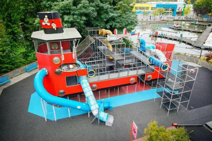 Rotes Spielschiff mit blauen R&ouml;hrenrutschen, Metallkletterger&uuml;sten und schwarzem Fallschutz, umgeben von B&auml;umen und weiteren Spielger&auml;ten
