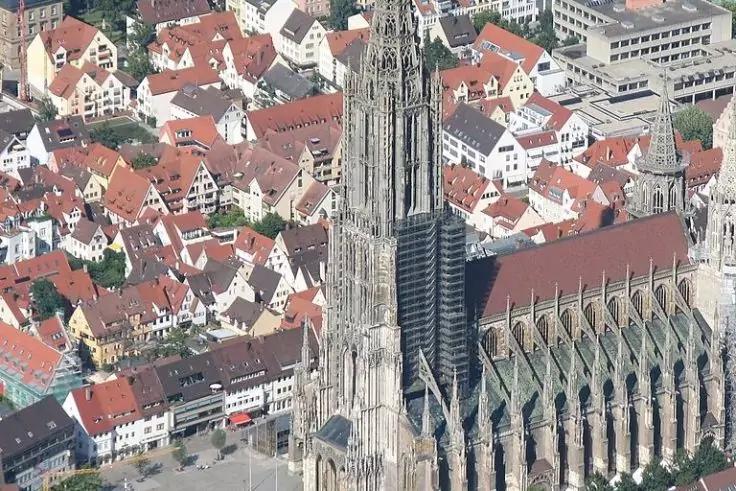 Luftaufnahme einer gro&szlig;en steinernen Kirche mit sehr hohem spitzem Turm, umgeben von roten und wei&szlig;en D&auml;chern