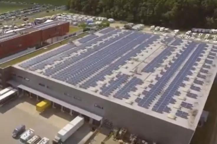 Draufsicht auf ein großes Industriegebäude mit Reihen von Solarpaneelen auf dem Flachdach, Ladebereich mit LKWs, geparkte Autos und Baumreihe im Hintergrund