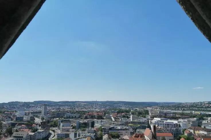 Blick &uuml;ber eine Stadt mit D&auml;chern, Geb&auml;uden und Kirchturm; im Hintergrund H&uuml;gel und blauer Himmel; oben links und rechts dunkle, dreieckige Rahmenteile