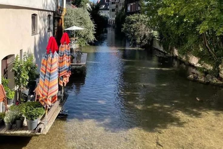 Schmaler Kanal zwischen H&auml;usern, linkes Ufer mit Pflanzgef&auml;&szlig;en und drei bunten, zusammengeklappten Sonnenschirmen; klares, flaches Wasser und B&auml;ume im Hintergrund