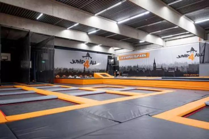 Mehrere rechteckige Trampoline mit orangefarbenen R&auml;ndern in einer Halle, Wandgrafik mit Schriftzug 'JUMP ALL' und Stadtsilhouette, Deckenleuchten