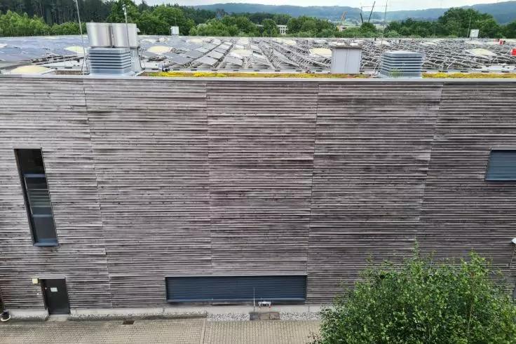 Gebäudefassade mit horizontalen Holzlatten, schmales horizontales Fenster unten in der Mitte, schmales vertikales Fenster links, Tür, Flachdach mit Lüftungsaufbauten und Büsche im Vordergrund