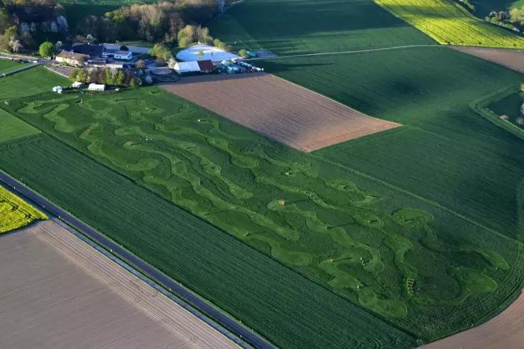 Luftaufnahme von Feldern mit geschwungenen Mustern in gr&uuml;ner Bepflanzung, angrenzenden braunen und gr&uuml;nen Feldern, einer Stra&szlig;e am Feldrand und Geb&auml;uden mit B&auml;umen am oberen Bildrand