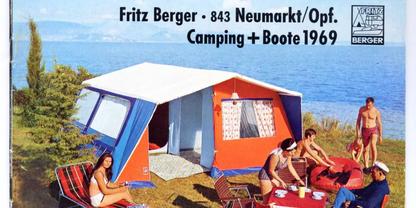 Rotes und blaues Campingzelt auf Gras am Ufer, f&uuml;nf Personen in Badebekleidung sitzen und stehen um einen kleinen Tisch mit Geschirr; See und bewaldete H&uuml;gel im Hintergrund