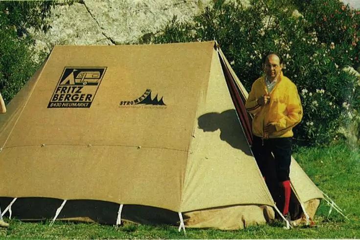 Beiges Zelt auf Gras mit aufgedrucktem 'Fritz Berger'-Logo und Bergsymbol; rechts steht eine Person in gelber Jacke am Zelteingang, Bäume im Hintergrund