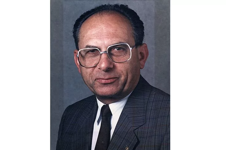 Porträt einer Person mit Brille, kurz zurückgekämmtem dunklem Haar, Nadelstreifenanzug, weißem Hemd, dunkler Krawatte und kleinem Ansteckpin vor grauem Hintergrund