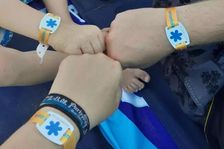 F&uuml;nf geballte H&auml;nde nah beieinander; Handgelenke mit orange-wei&szlig;en Armb&auml;ndern mit blauem sternf&ouml;rmigem Symbol; blauer Stoff im Hintergrund
