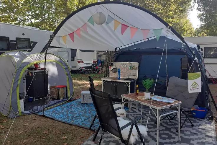 Halbrunde wei&szlig;e Campingmarkise mit bunten Wimpeln, davor schwarzer Klappstuhl, wei&szlig;er Campingtisch mit kleiner Topfpflanze und Zeitschrift, zwei gemusterte Bodenmatten, K&uuml;chenbox mit Wasserhahn, mehrere Wohnwagen im Hintergrund