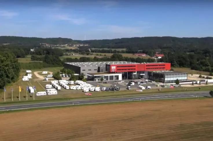 Luftaufnahme eines großen roten Gebäudes neben einem Parkplatz mit mehreren weißen Wohnwagen und Autos, eine Straße im Vordergrund, Felder und bewaldete Hügel im Hintergrund unter blauem Himmel