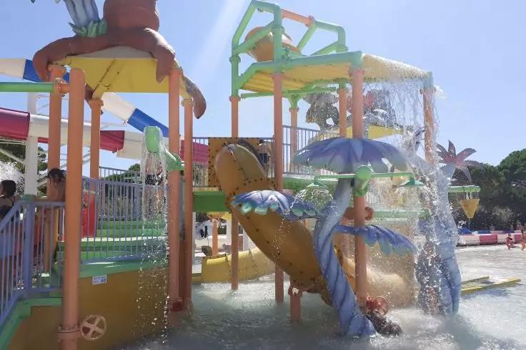 Bunte Wasserspielanlage mit mehrstufiger Struktur, Rutschen, Wasser, das aus kippenden Eimern und schirmf&ouml;rmigen Spr&uuml;hk&ouml;pfen flie&szlig;t, und flachem Becken