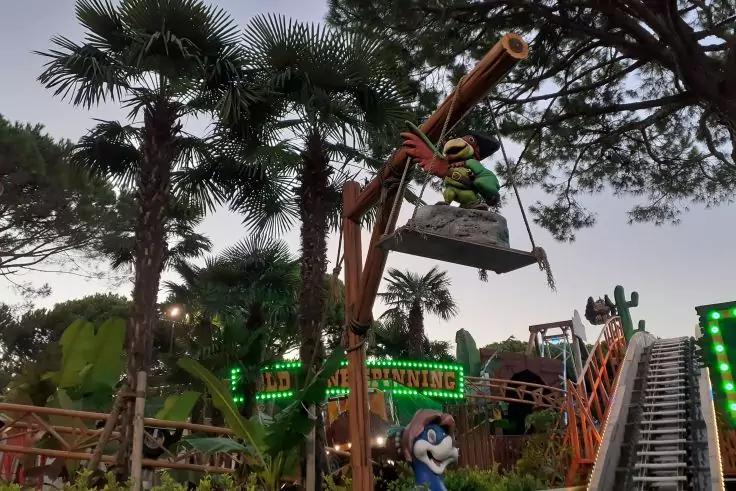 Person in einem h&auml;ngenden Metallkorb an einem h&ouml;lzernen Ausleger; im Hintergrund Palmen, rechts Schiene mit Treppe, unten eine blaue Cartoon-Figur, Abendhimmel