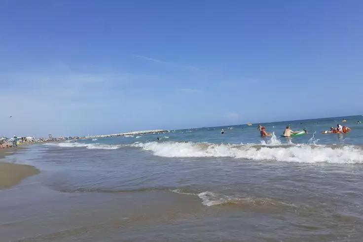 Sandiger Strand, Wellen am Ufer, mehrere Personen auf kleinen Booten oder Boards im Wasser, blauer Himmel