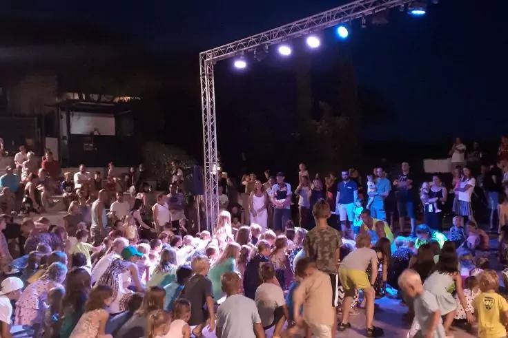 Menschenmenge vor einer beleuchteten Metallb&uuml;hnenkonstruktion bei Nacht, einige sitzen, andere stehen