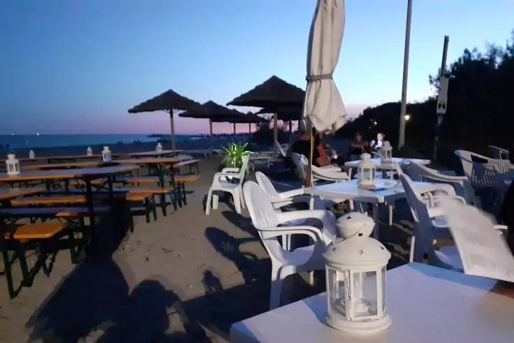 Tische und wei&szlig;e Plastikst&uuml;hle auf sandigem Boden, wei&szlig;e Laterne auf einem Tisch im Vordergrund, Sonnenschirme, Meer am Horizont, einige Personen im Hintergrund