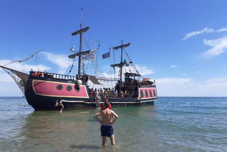 Gro&szlig;es Segelschiff mit schwarzem und pinkem Rumpf nahe der K&uuml;ste im flachen Wasser, mehrere Masten und Takelage, Menschen an Bord und im Wasser, Mann im Vordergrund mit blauer Badehose, klarer Himmel mit wenigen Wolken