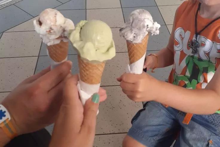 Drei H&auml;nde halten Waffelh&ouml;rnchen mit jeweils einer Kugel Eis; gefliester Boden; Teile eines Kindes mit orangefarbenem Oberteil und Shorts sichtbar